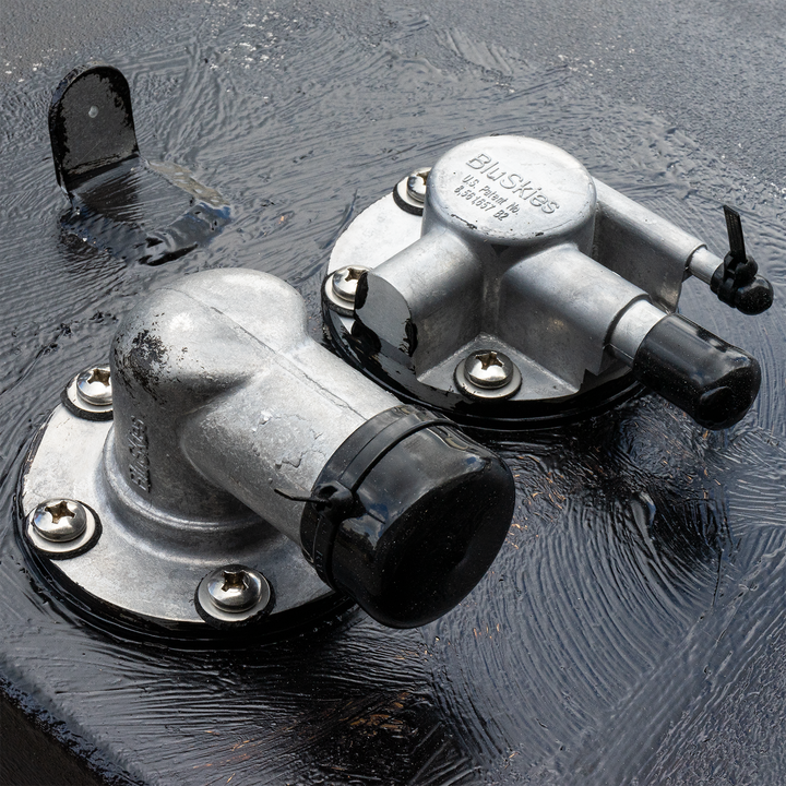 Close-up view of two silver fuel tank fittings on a black surface, featuring a fuel fill cap and a vent, with visible screws and a slightly wet appearance
