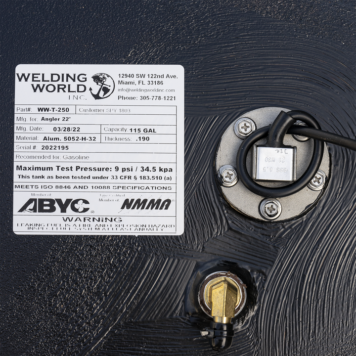 Close-up of a black fuel tank surface with a white product label from Welding World Inc. detailing specifications like part number WW-T-250, 115 gal capacity, and maximum test pressure, alongside a fuel level sender with wiring and a brass fitting