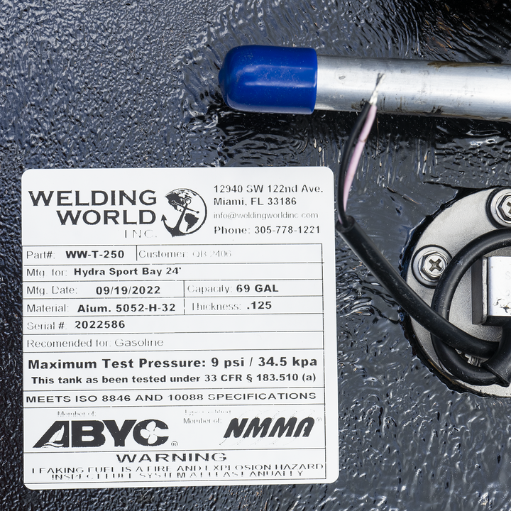 A close-up of a white manufacturer's label on a heavily textured black fuel tank. A metal tube with a blue cap is visible above the label, and a round, silver fuel sending unit is partially seen to the right. The "Welding World, Inc." label identifies the tank is for a "Hydra Sport Bay 24'" with a 69-gallon capacity, made on September 19, 2022. The label also features ABYC and NMMA compliance logos and a safety warning