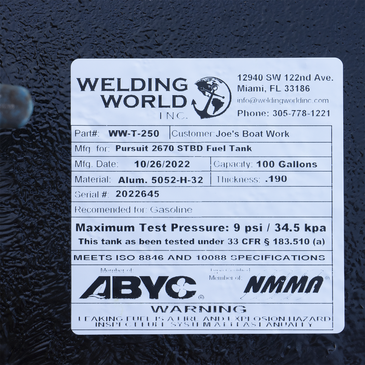 A close-up photograph of a white manufacturer's label on a black, textured surface. The label from "Welding World, Inc." identifies the item as a "Pursuit 2670 STBD Fuel Tank" with a 100-gallon capacity, manufactured on October 26, 2022. Compliance logos for ABYC and NMMA are displayed above a safety warning at the bottom of the label