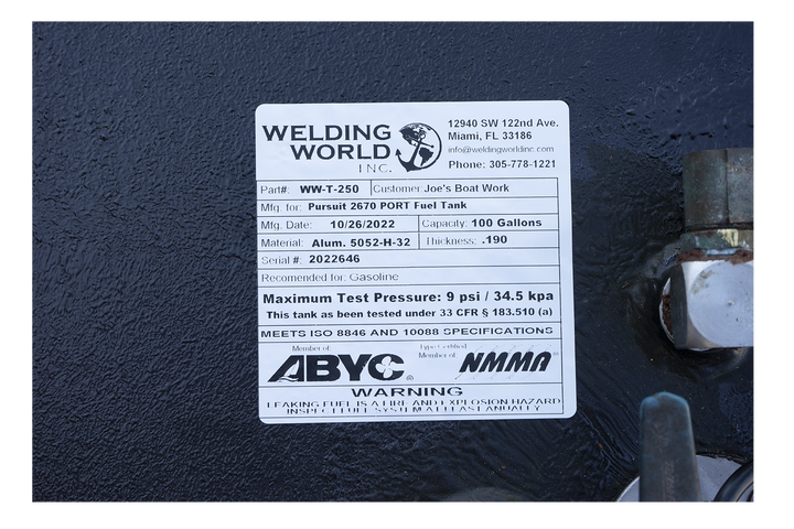 A close-up photograph of a white manufacturer's label on a black, textured surface. The label from "Welding World, Inc." identifies the item as a "Pursuit 2670 STBD Fuel Tank" with a 100-gallon capacity, manufactured on October 26, 2022. Compliance logos for ABYC and NMMA are displayed above a safety warning at the bottom of the label.