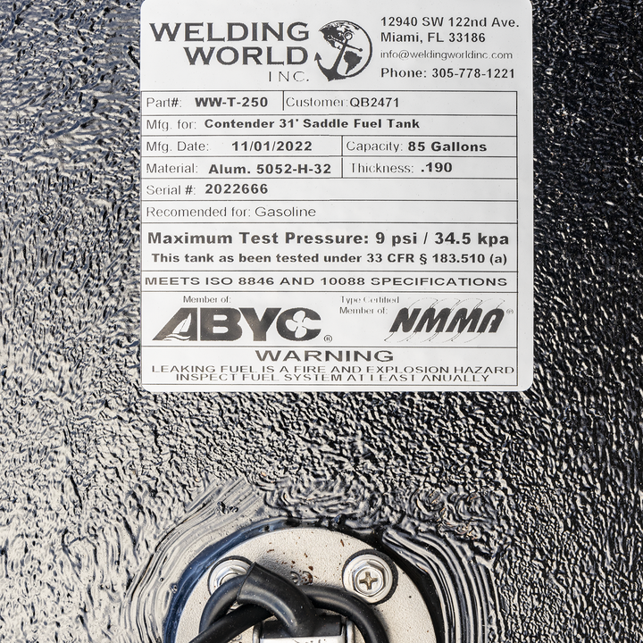 A close-up view showing a white manufacturer's label positioned above a round, silver fuel sending unit on a heavily textured black surface. The "Welding World, Inc." label identifies the product as a "Contender 31' Saddle Fuel Tank" with an 85-gallon capacity, made on November 1, 2022. Below, the sending unit is recessed into the tank with a black wire emerging from its center. The label also displays ABYC and NMMA compliance logos