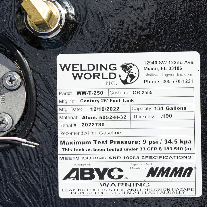 A close-up photo of a white rectangular label on a black metal surface. The label is from Welding World Inc., located at 12940 SW 122nd Ave., Miami, FL 33186, with phone number 305-778-1221. The label contains detailed specifications for a Century 26' Fuel Tank, manufactured on 12/19/2022. It lists the part number WW-T-250, customer QB 2555, material Alum. 5052-H-32, thickness .190, and serial number 2022780. The tank has a capacity of 134 gallons and is recommended for gasoline