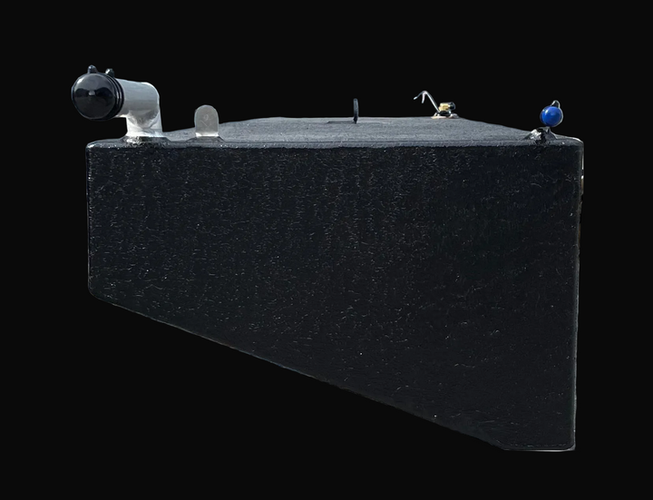 A close-up, end-on profile photograph of a long black marine fuel tank, highlighting its rectangular cross-section and textured finish. The image shows the installation of a silver-toned fill neck and a smaller blue-capped vent fitting on the top edge, as well as a central silver lifting eye and brass fittings in the background.