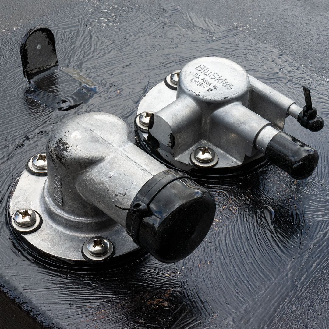 Close-up view of two silver fuel tank fittings on a black surface, featuring a fuel fill cap and a vent, with visible screws and a slightly wet appearance