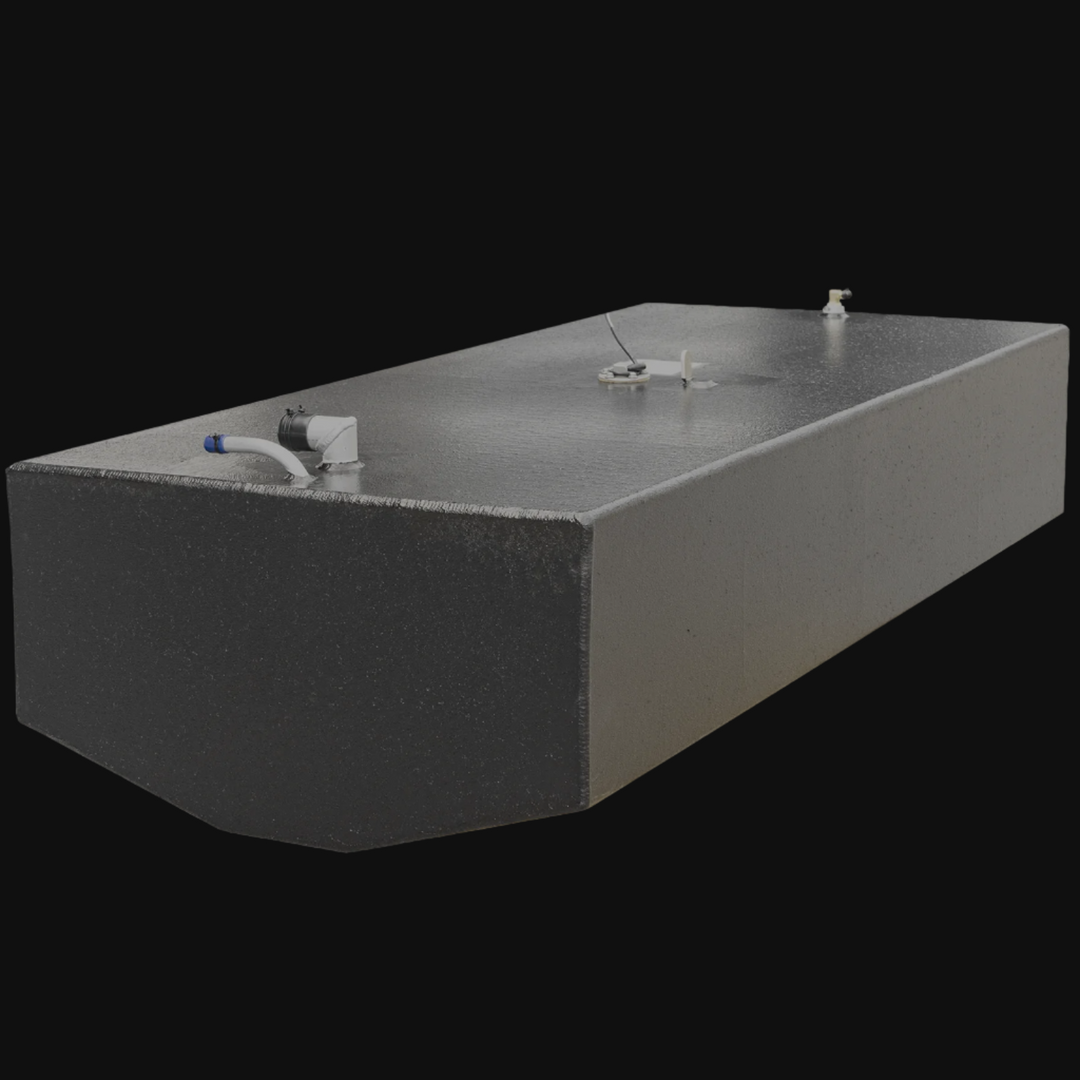 A photo of a large, rectangular metallic fuel tank with a black textured finish on the front and a lighter finish on the sides. The tank has several fittings and components mounted on the top surface, including pipes, connectors, and caps. The background is black