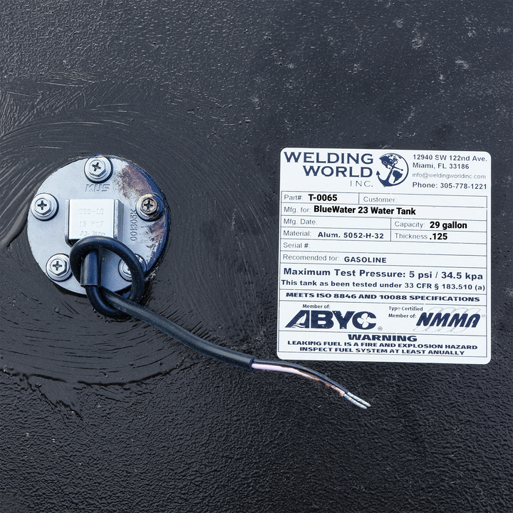 Close-up photograph of a Welding World Inc. manufacturing label for a BlueWater 23 tank, specifying a 29-gallon capacity, .125" thickness, and 5052-H32 aluminum construction, shown next to a KUS unit on a black epoxy finish.