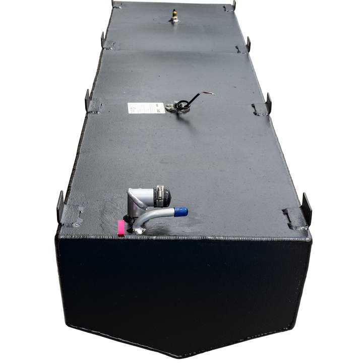 A long, black, rectangular fuel tank with a textured surface and multiple metal brackets along its top edges. Various fittings are visible on the top surface, including a fuel sender with wiring, a small brass fitting, and an L-shaped hose connection with a blue cap and a pink marker