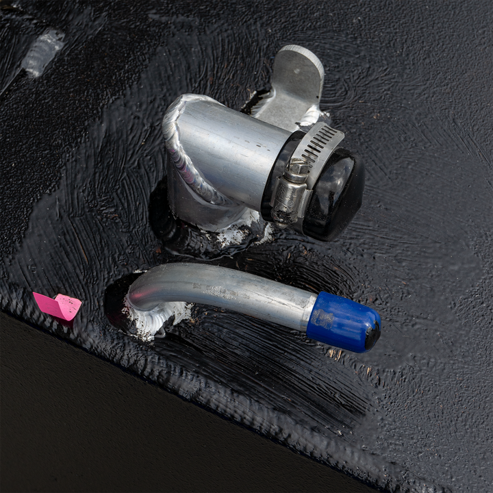 A close-up shot of the top surface of a black, textured fuel tank, showing various metallic and plastic fittings. This includes a curved silver pipe with a blue cap, an L-shaped silver pipe with a black cap and hose clamp, and a small, flat metal piece, all visibly welded to the tank. A pink, small flag-like object is also visible
