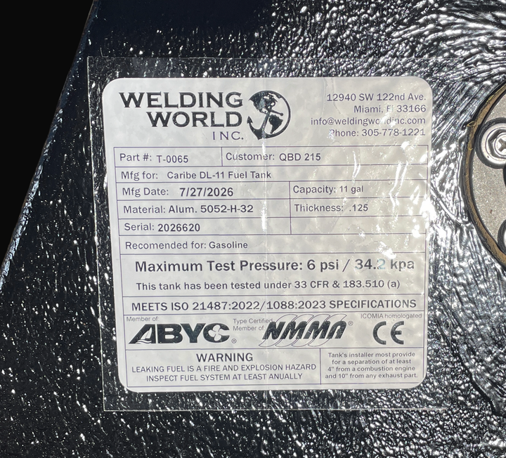 A macro top-down view of the official manufacturer certification tag on a physical, finished black encapsulated Caribe DL-11 OEM replacement marine fuel tank (11 gal 2016) from file image_aeb39a.jpg. Adhered to the textured black TarGard epoxy coating, the weather-resistant specification label from Welding World Inc. (Miami, FL) lists part number T-0065, heavy-gauge Alum. 5052-H-32 material, .125 thickness, an 11 gal capacity, gasoline recommendation, 6 psi / 34.2 kpa maximum test pressure under 33 CFR & 18