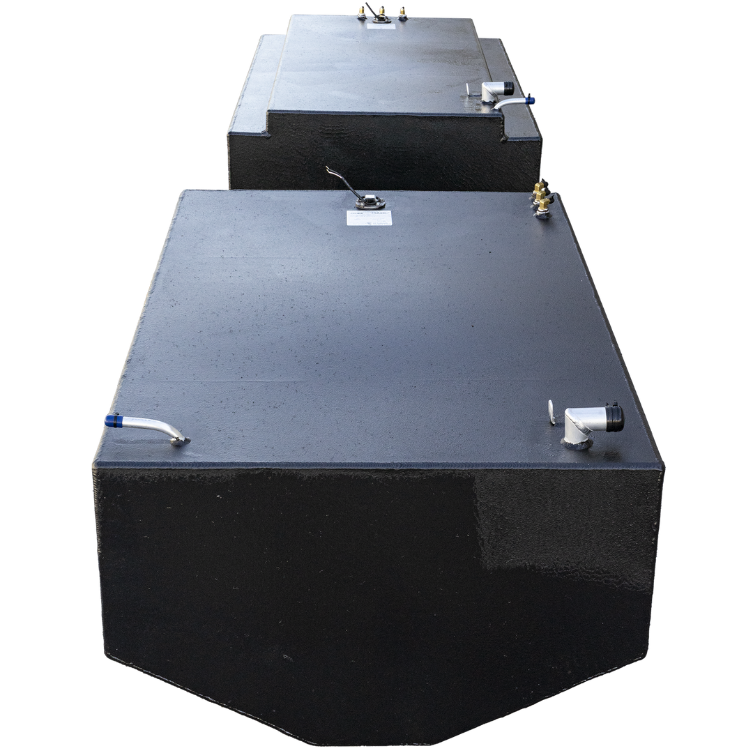 Two black, rectangular fuel tanks are positioned one behind the other. The front tank has a slightly rounded front edge and a variety of fittings on its top, including a white L-shaped pipe with a blue cap on the left and a silver L-shaped pipe on the right. The back tank has a stepped design and also features fittings on its top surface. Both tanks have a textured finish