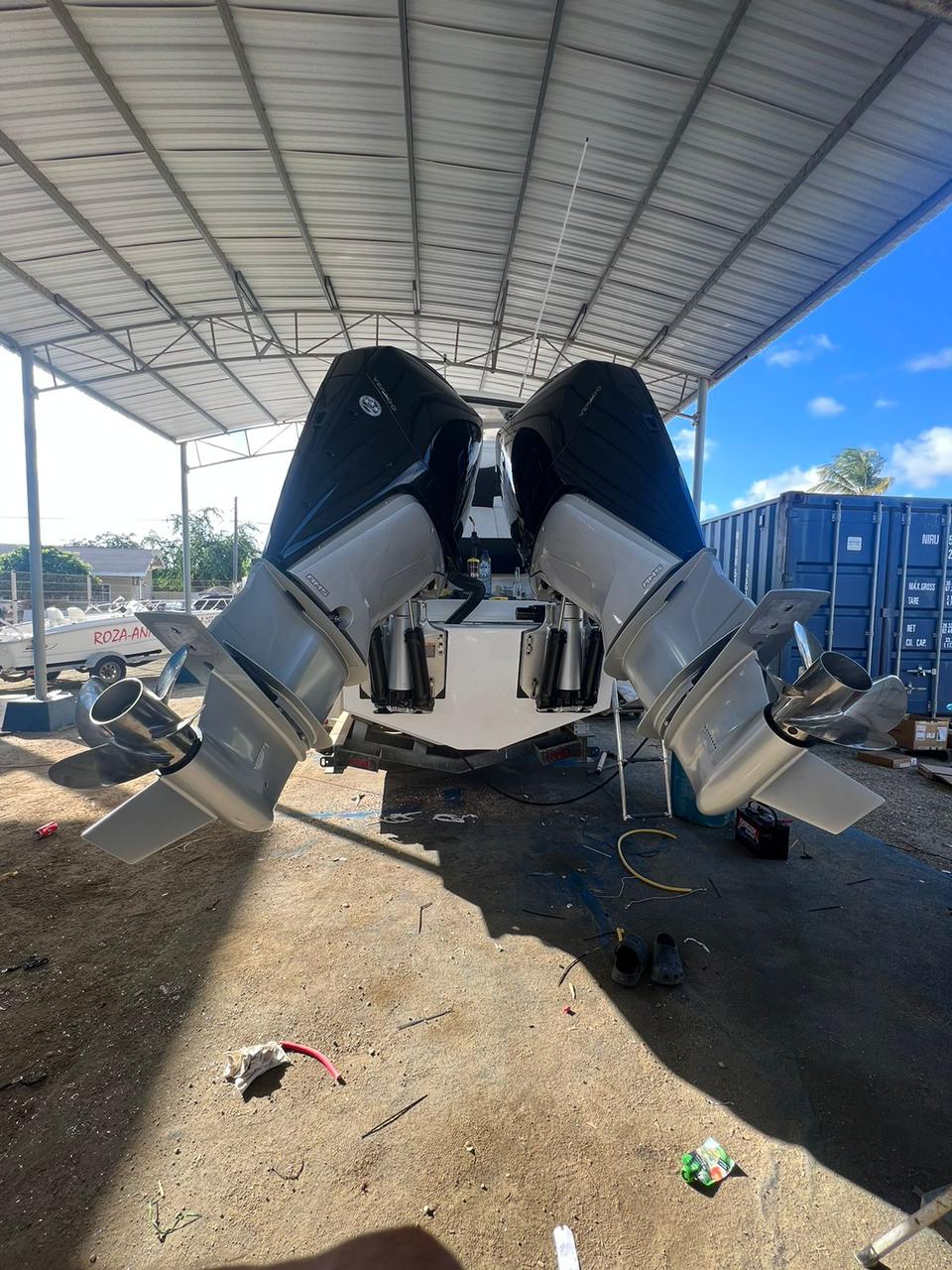Alt text: The back view of a boat with two large outboard engines mounted on the stern, inside a covered area with a metal roof. The engines have black and silver casings, and the boat is supported on a stand. There are various tools, cables, and equipment on the ground around the boat, with a blue shipping container and other boats visible in the background