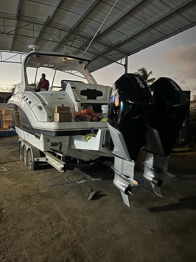 A white boat with two large black outboard engines mounted at the stern, parked underneath a metal roof structure. The boat has various equipment and boxes on board, and a person is standing in the background. The sky is partly cloudy with some palm trees visible