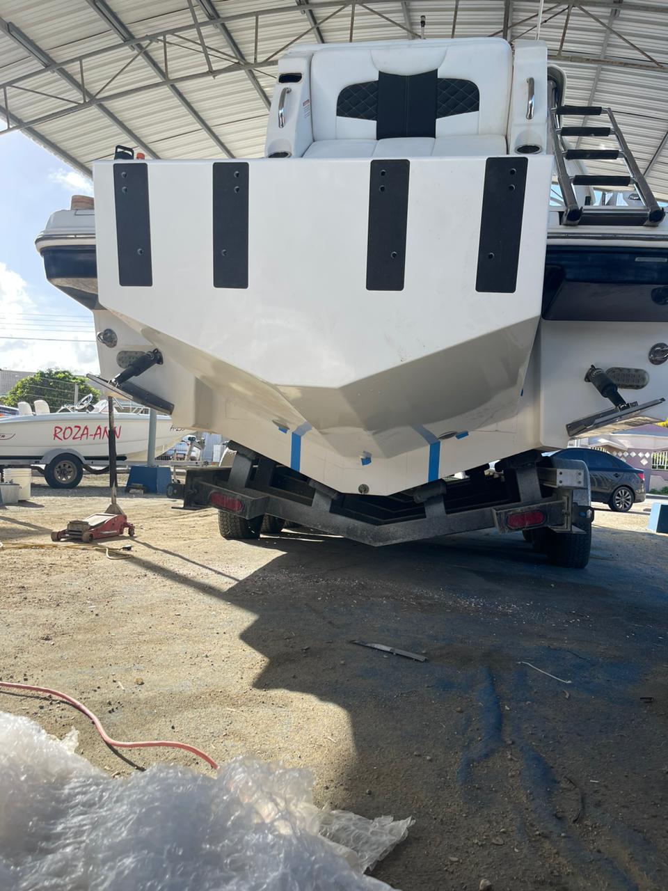 The front view of a white Chaparral 327 SSX boat with a custom high flotation twin outboard engine bracket, parked on a trailer under a metal canopy. The boat's bow features black accents and a seating area, and there are other boats and vehicles visible in the background. The boat is supported by a black trailer with red taillights, and the ground is a dirt surface