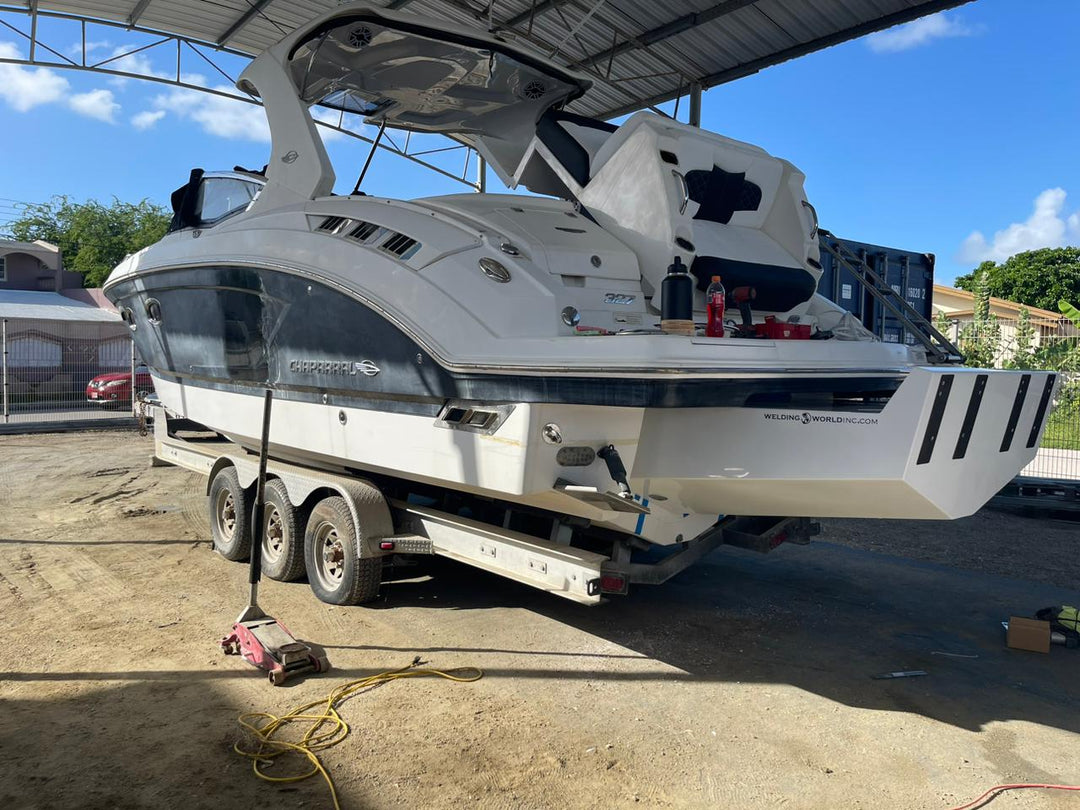 A white Chaparral 327 SSX boat with a custom high flotation twin outboard engine bracket, parked on a trailer under a metal roof structure. The boat is in an outdoor storage area with some tools and equipment around, and the sky is clear with a few clouds