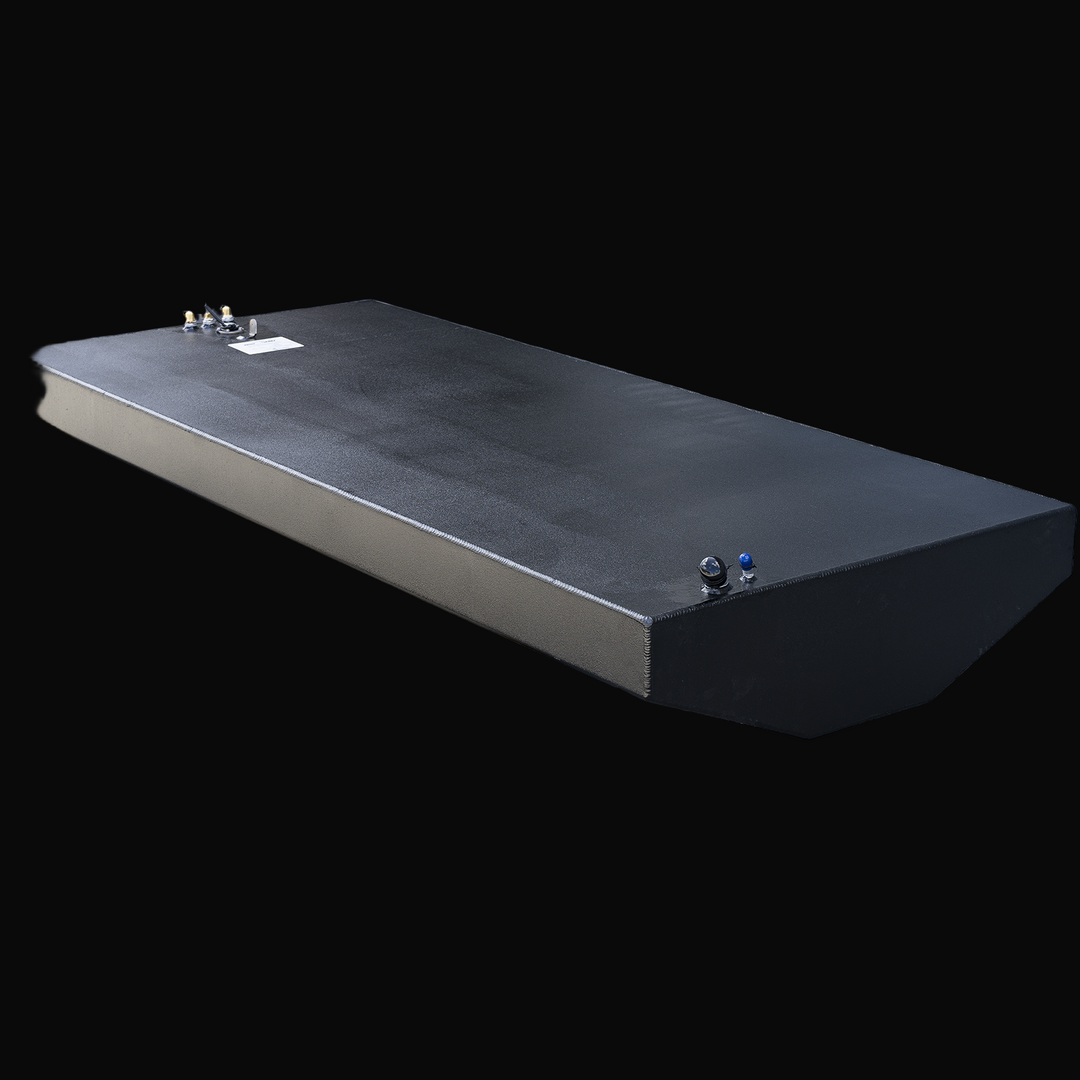 Long rectangular black fuel tank with beveled front edge and straight vertical sides, viewed from the top left front corner. The top surface has multiple fittings and labels near the front left corner and one set near the back right corner. The tank has visible weld seams along the edges and a textured finish. Background is transparent