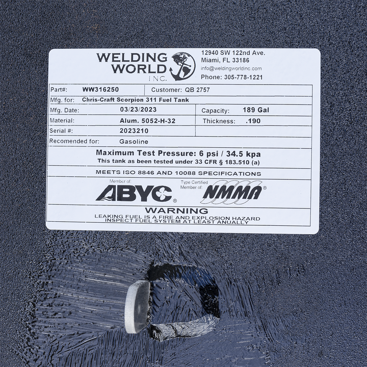 Close-up view of a white rectangular label on a textured black surface. The label is from Welding World Inc., located at 12940 SW 122nd Ave, Miami, FL 33186, with contact info and phone number 305-778-1221. It shows part number WW316250, customer QB 2757, manufacturing for Chris-Craft Scorpion 311 Fuel Tank, manufacturing date 03/23/2023, capacity 189 gallons, material Aluminum 5052-H-32, thickness 0.190, serial number 2023210, recommended for gasoline