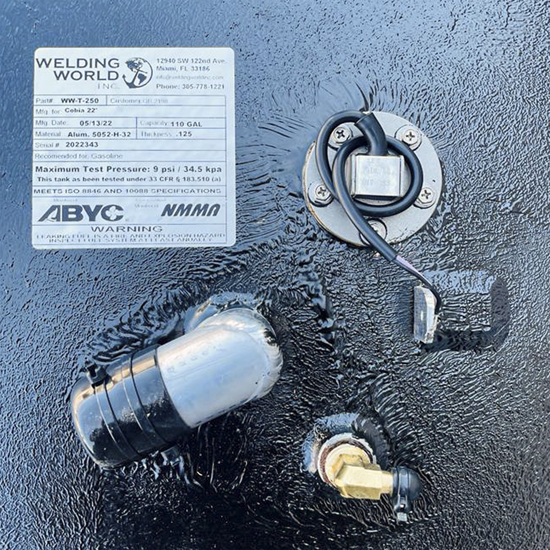 A white product label from "WELDING WORLD INC." is adhered to a textured black surface, likely a fuel tank. The label lists product specifications, including Part# WW-T-250, Mfg. for Cobia 22', Mfg. Date 05/13/22, Capacity 110 GAL, Material Alum. 5052-H-32, Thickness .125, and Serial # 2022343. It is recommended for Gasoline, has a maximum test pressure of 9 psi / 34.5 kpa, and meets ISO 8846 and 10088 specifications. Below the label, various fittings and pipes are visible on the black surface