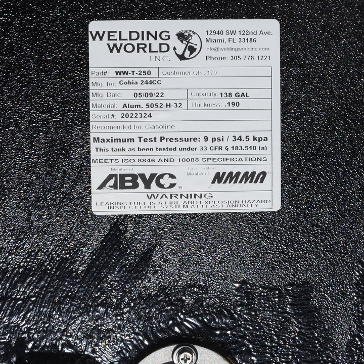 A close-up of a white product label from "Welding World Inc." on a textured black surface. The label provides specifications for a fuel tank, including Part#, Mfg. for, Mfg. Date, Material, Serial #, Capacity, and Thickness. It also states the maximum test pressure, certification details, and a warning about leaking fuel
