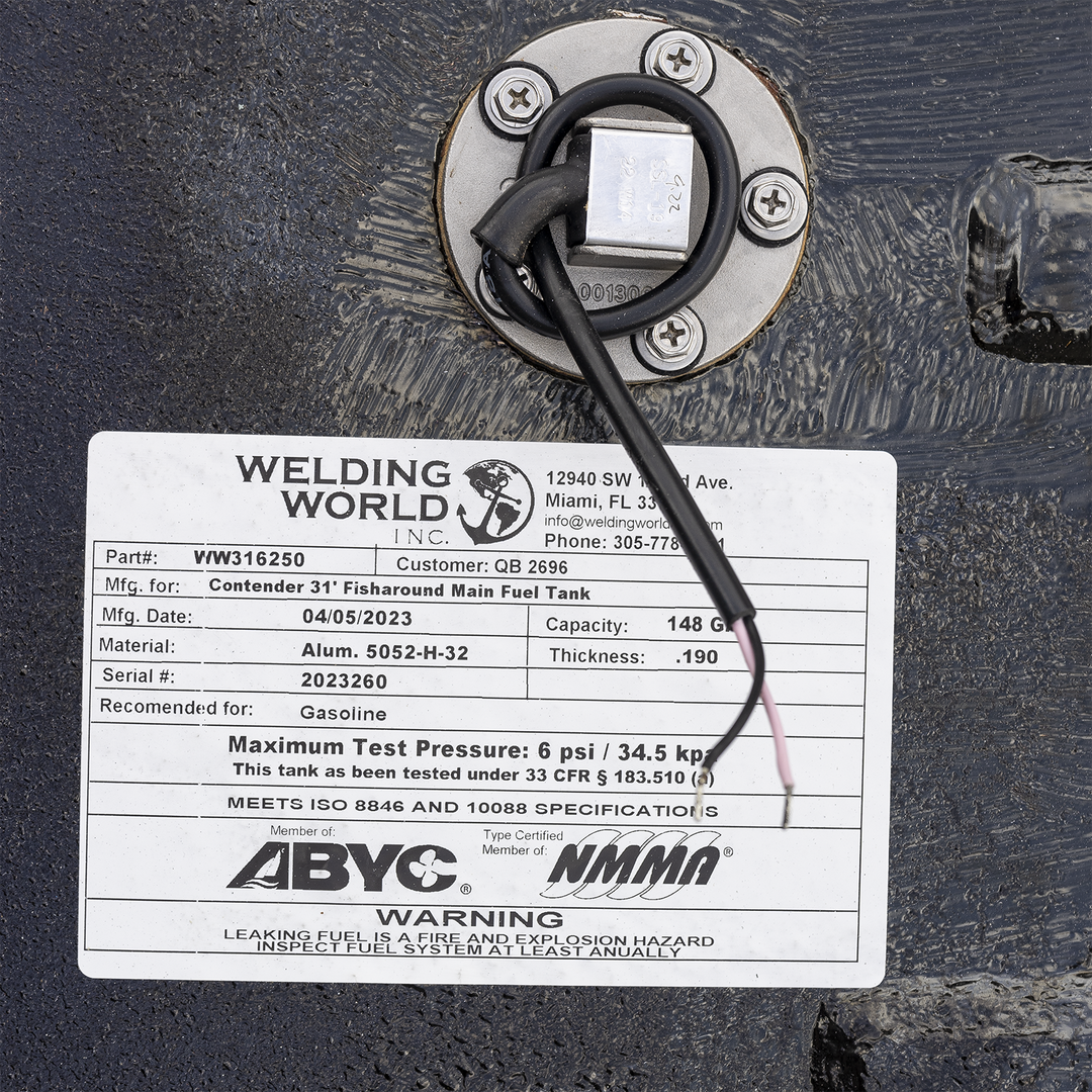 Close-up of a white label attached to a black textured metal surface. The label shows Welding World Inc. company info with address 12940 SW 122nd Ave., Miami, FL 33186 and phone number 305-778-1221. It lists part number VW316250 and customer QB 2696. The label is for a Contender 31' Fisharound Main Fuel Tank, manufactured on 04/05/2023. Capacity is 148 gallons, made of Aluminum 5052-H-32 with thickness 0.190. Serial number 2023260. Recommended for gasoline. Maximum test pressure is 6 psi / 34.5 kpa