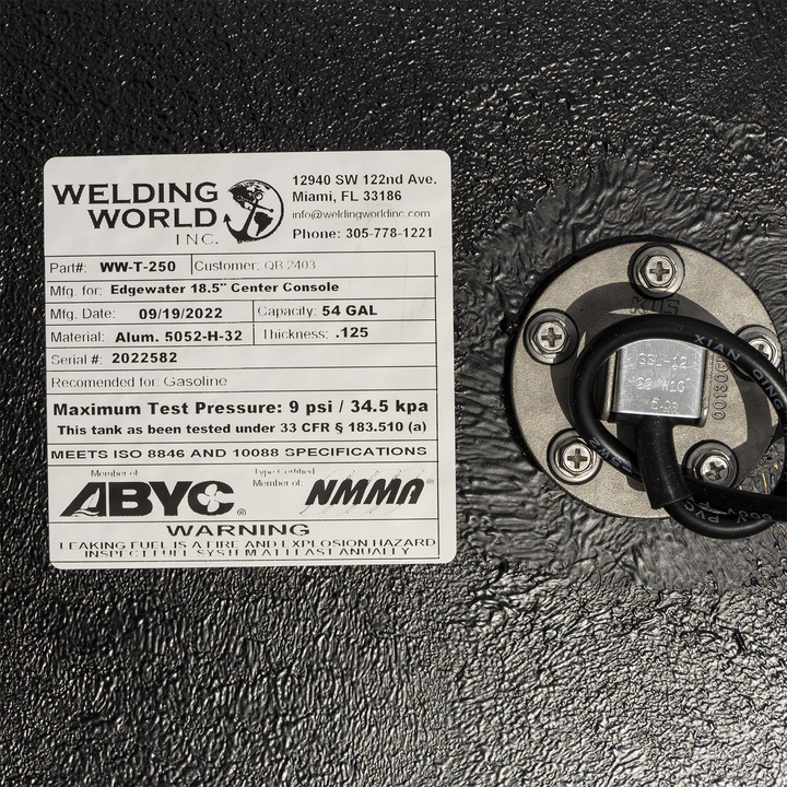 A close-up view of a manufacturer's label and a round fuel sending unit on a heavily textured black surface. On the left, the white "Welding World, Inc." label specifies the tank is for an "Edgewater 18.5" Center Console" with a 54-gallon capacity, made on September 19, 2022. On the right, the silver fuel sending unit is secured with four screws, with a black wire emerging from its center. The label also features ABYC and NMMA compliance logos