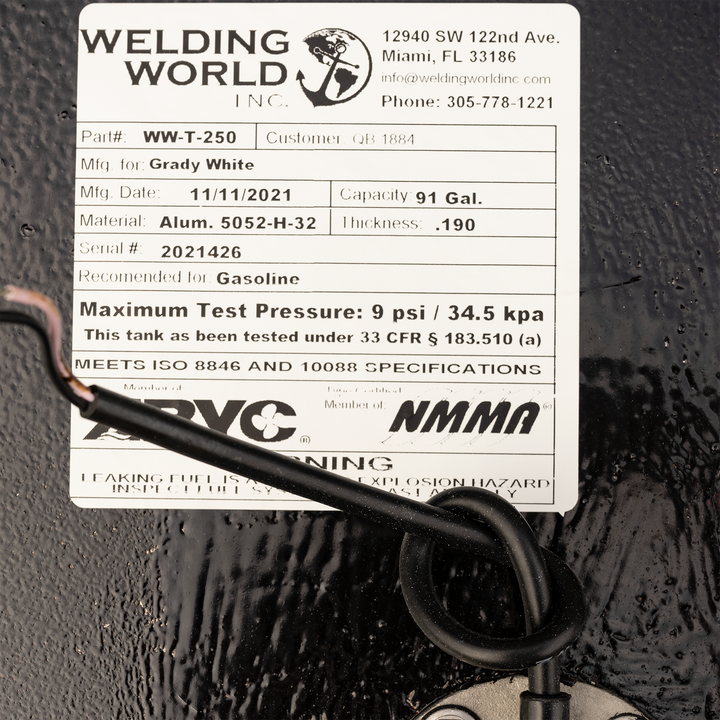 A close-up of a white product label from "WELDING WORLD INC." on a black, textured surface. The label details a fuel tank for Grady White, Part# WW-T-250, manufactured on 11/11/2021, with a capacity of 91 gallons, made from Alum. 5052-H-32 with a thickness of .190, Serial # 2021426. It is recommended for Gasoline and has a maximum test pressure of 9 psi / 34.5 kpa. The label also states it meets ISO 8846 and 10088 specifications and is a member of ABYC and NMMA, with a warning about leaking fuel
