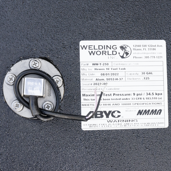 A close-up view of a product label from 'Welding World Inc.' affixed to a textured black surface. The label provides detailed specifications for a 'Hewes 16' Fuel Tank,' including Part # WW-T-250, Mfg. Date 08/01/2022, Capacity 30 GAL, Material Alum. 5052-H-32, Thickness .125, and Serial # 2022290. It also states the tank is recommended for Gasoline, has a Maximum Test Pressure of 9 psi / 34.5 kpa, and meets ISO 8846 and 10088 specifications. Logos for ABYC and NMMA are present