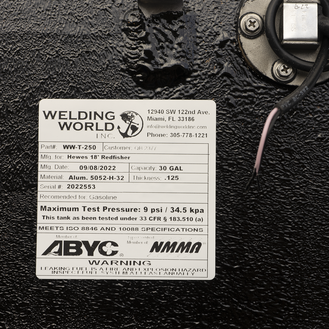 A close-up view of a product label from 'Welding World Inc.' attached to a black, textured surface. The label provides detailed specifications for a fuel tank manufactured for 'Hewes 18' Redfisher', including Part # WW-T-250, Mfg. Date 09/08/2022, Capacity 30 GAL, Material Alum. 5052-H-32, Thickness .125, and Serial # 2022553. It also states the tank is recommended for Gasoline, has a Maximum Test Pressure of 9 psi / 34.5 kpa, and meets ISO 8846 and 10088 specifications