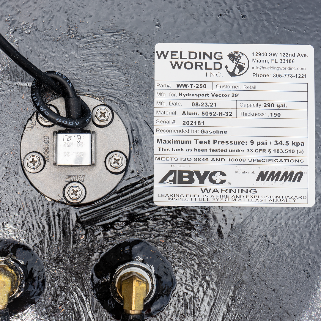 A white label on a black fuel tank, indicating it's a 290-gallon fuel tank for a Hydrasport Vector 29', manufactured by Welding World Inc. on 08/23/21. The label specifies part number WW-T-250, material Alum. 5052-H-32 with .190 thickness, serial number 202181, recommended for gasoline, with a maximum test pressure of 9 psi / 34.5 kpa. It also states compliance with ISO 8846 and 10088 specifications and certifications by ABYC and NMMA, along with a warning about fuel leaks. Various metal fittings
