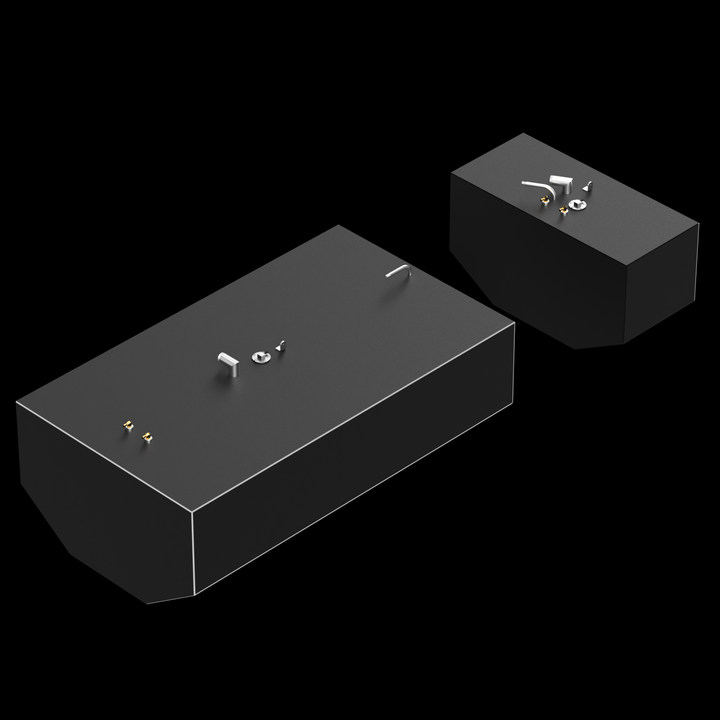 Two dark grey, rectangular fuel tanks, of different sizes, are shown against a black background. The larger tank is in the foreground and has several metallic fittings on its top surface, including two brass-colored connectors and a larger silver-colored component. The smaller tank is in the background and also has various metallic fittings on its top surface. Both tanks have a white outline along their top edges