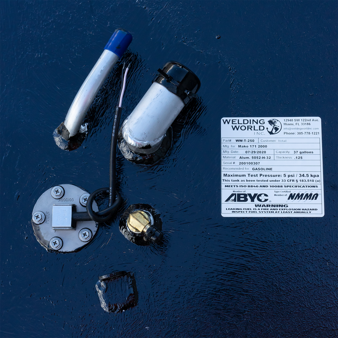 A top-down close-up of various fuel fittings and a manufacturer's label on a heavily textured black surface. The components include a round silver fuel sending unit, a large brass fitting, and two angled pipes with blue and black caps. The white "Welding World, Inc." label identifies the tank for a "Mako 171 2000" with a 37-gallon capacity, made on July 29, 2020, and displays ABYC and NMMA compliance logos