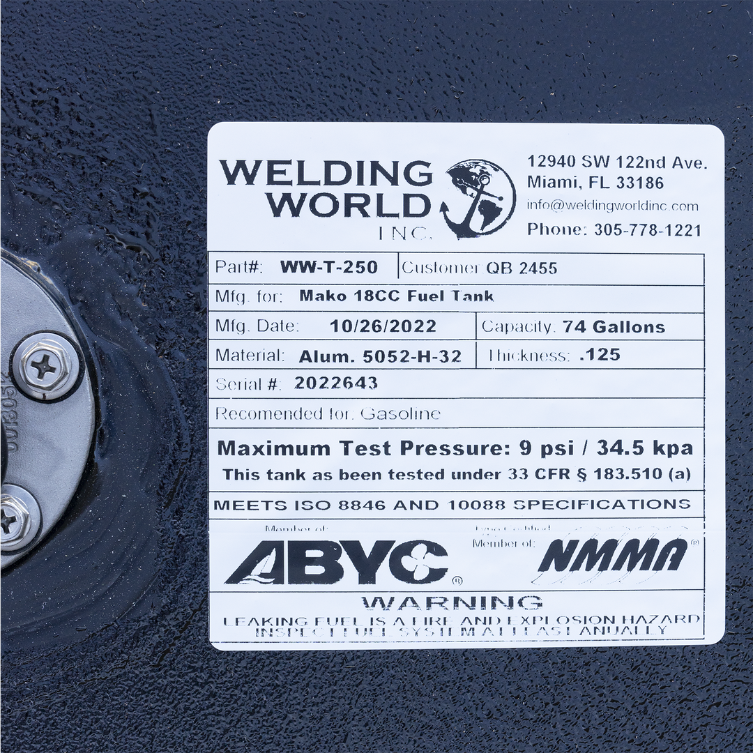 A close-up of a white manufacturer's label on a black textured fuel tank. To the left of the label, the silver flange of a fuel sending unit is partially visible. The "Welding World, Inc." label identifies the product as a "Mako 18CC Fuel Tank" and lists specifications including a 74-gallon capacity, an aluminum construction, and a manufacturing date of October 26, 2022. Compliance logos for ABYC and NMMA and a safety warning are at the bottom