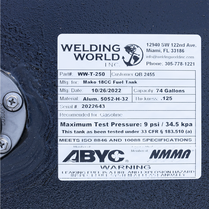 A close-up of a white manufacturer's label on a black textured fuel tank. To the left of the label, the silver flange of a fuel sending unit is partially visible. The "Welding World, Inc." label identifies the product as a "Mako 18CC Fuel Tank" and lists specifications including a 74-gallon capacity, an aluminum construction, and a manufacturing date of October 26, 2022. Compliance logos for ABYC and NMMA and a safety warning are at the bottom