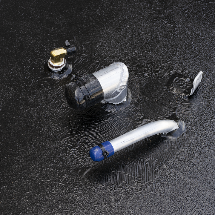 A close-up view of various metal fittings on a textured black surface, appearing wet or oily. The fittings include a small brass elbow connector, a larger silver-colored elbow with a black cap, and a silver pipe with a blue cap and a black zip tie. There are visible ripples and reflections on the surface around the fittings