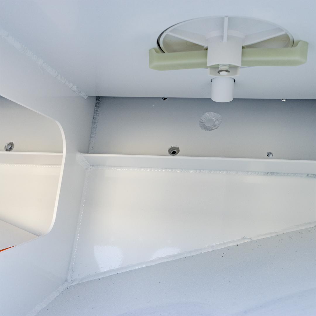 Close-up view of the interior structure of a boat, showing a white surface with a mounted circular component at the top, a green support arm, and several screw holes and welds visible on the metal surface