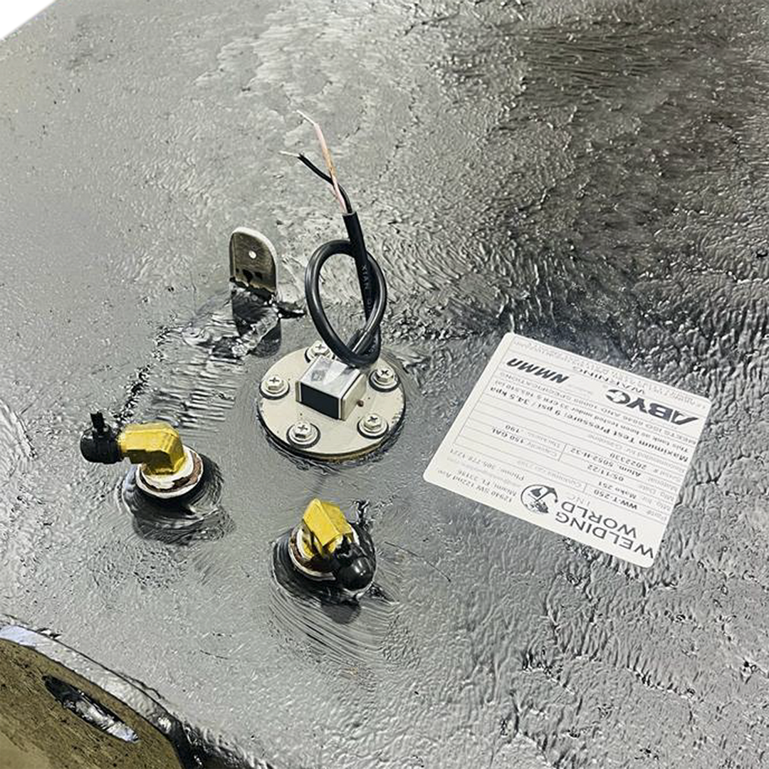 An overhead close-up view of a textured dark grey surface, likely a fuel tank, shows a white product label from "WELDING WORLD INC." detailing product specifications. Below and to the left of the label, two brass fittings with black caps are visible. A circular metallic plate secured by screws with a black wire extending from its center is also present