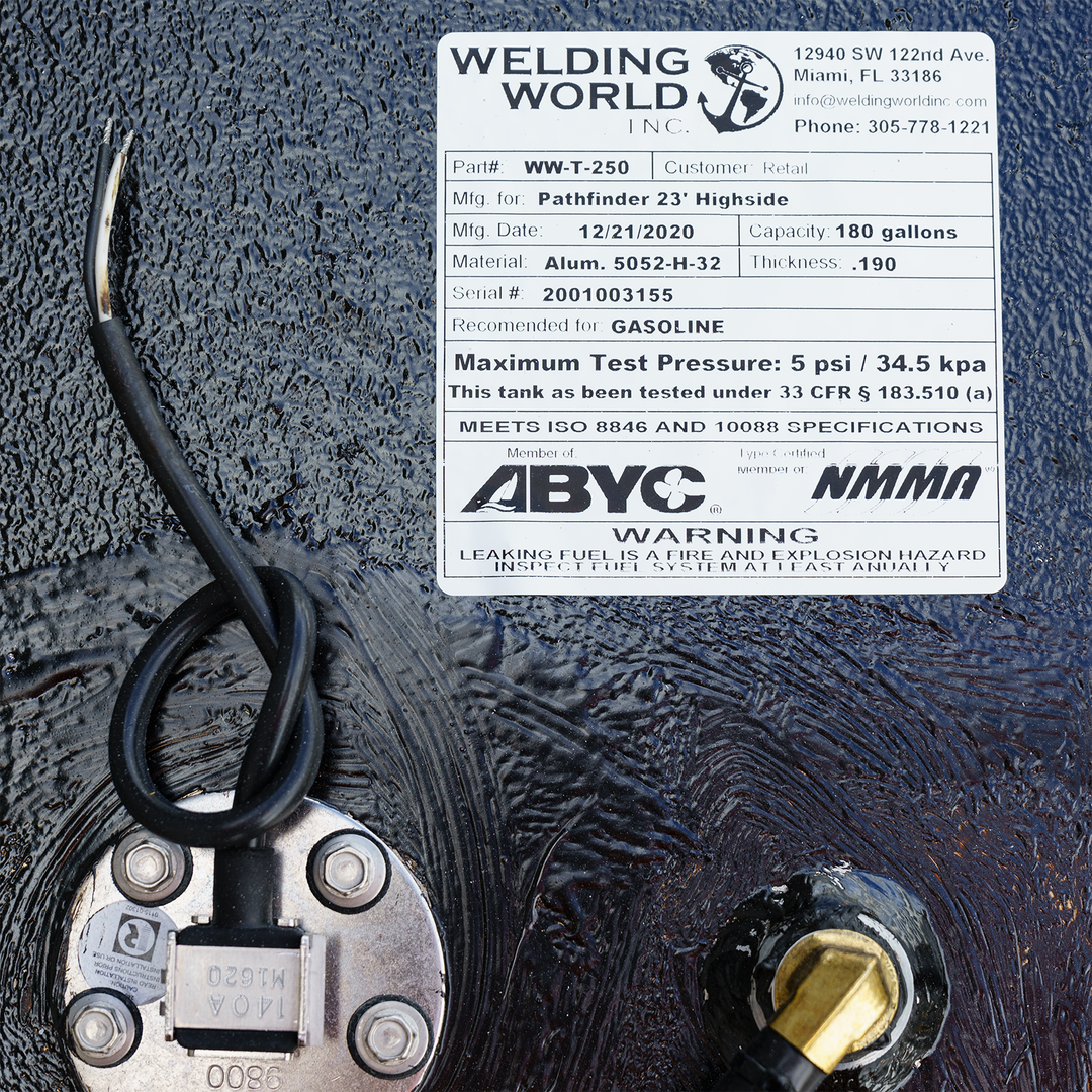 A close-up image of a specification label from Welding World Inc., detailing the fuel tank's part number (WW-T-250), manufacturing data for the Pathfinder 23' Highside with 180-gallon capacity, material specifications, and safety warnings about leaking fuel hazards. The label includes a serial number, testing pressure information, and compliance certifications, along with two wires connected to the tank