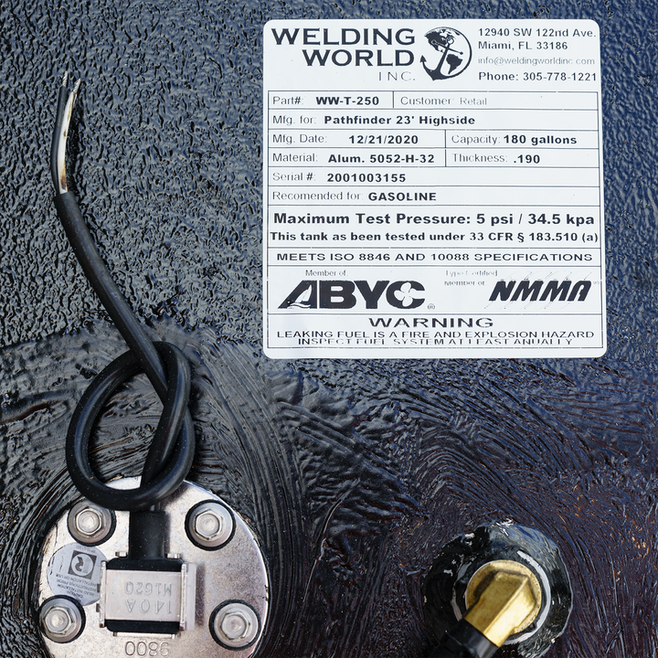 A close-up image of a specification label from Welding World Inc., detailing the fuel tank's part number (WW-T-250), manufacturing data for the Pathfinder 23' Highside with 180-gallon capacity, material specifications, and safety warnings about leaking fuel hazards. The label includes a serial number, testing pressure information, and compliance certifications, along with two wires connected to the tank