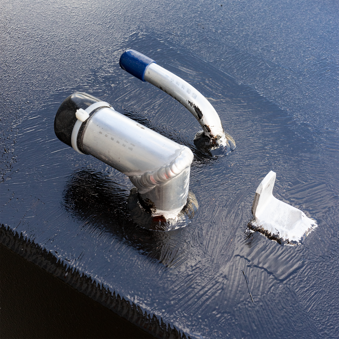 A close-up view of a black, textured surface, possibly a fuel tank, adorned with several metal components. These include two silver-colored pipes, one with a black cap and white zip tie, and the other with a blue cap, and a smaller, L-shaped silver bracket, all appearing to be welded or sealed to the surfac