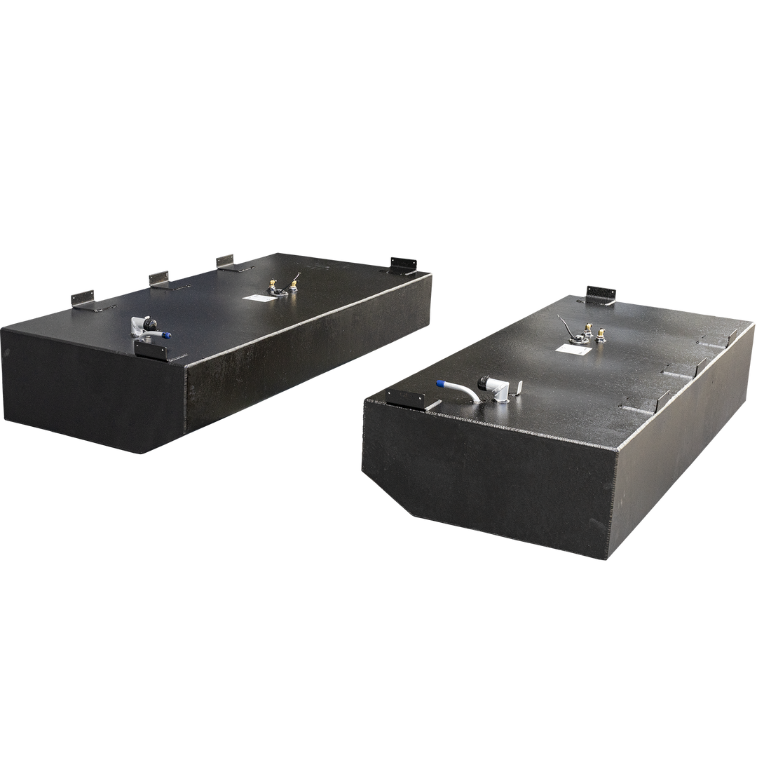 Two black, rectangular fuel tanks with textured surfaces are shown. The tank in the foreground has a rounded front edge and a white pipe with a blue tip on its top right. The tank in the background is fully rectangular and has a circular fitting with several brass-colored components on its top left. Both tanks feature multiple metal brackets attached to their top edges