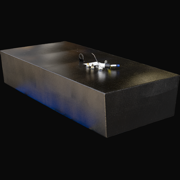 A large black rectangular fuel tank with a slightly beveled front bottom edge, viewed from the top front-left angle. The top surface features multiple fittings and connectors, including a circular flange, fuel sender unit, and wiring. The tank has a textured black finish and a subtle blue reflection along the left side