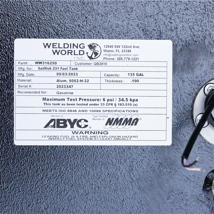A white rectangular label affixed to a textured black surface, showing specifications for a Sailfish 231 Fuel Tank. The label includes the Welding World Inc. logo with an anchor graphic, contact information for the company in Miami, FL, and various details such as part number WW316250, customer QB2815, manufacturing date of 05/03/2023, material Aluminum 5052-H-32, thickness .190, serial number 2023347, and a capacity of 135 gallons. It also states the tank is recommended for gasoline