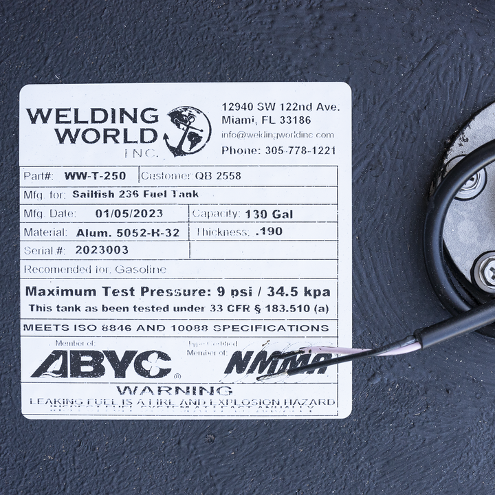 A close-up photo of a white label on a textured black surface showing product information from Welding World Inc. The label indicates the part number WW-T-250 for a Sailfish 236 Fuel Tank manufactured on 01/05/2023. The tank has a capacity of 130 gallons and is made of aluminum 5052-K-32 with a thickness of .190 inches. The serial number is 2023003, and the tank is recommended for gasoline use. The label also notes a maximum test pressure of 9 psi / 34.5 kpa and compliance with ISO 8846 and 10088