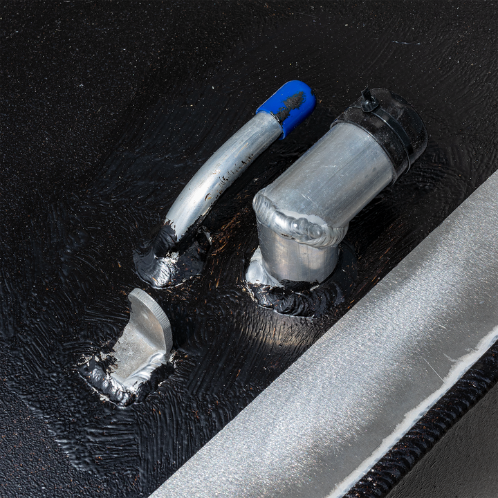 A close-up shot of the top surface of a black, textured fuel tank, showing various metallic fittings and components. This includes a curved silver pipe with a blue cap, an L-shaped silver pipe with a black cap, and a small, flat, angled metal piece. All components are visibly welded to the tank surface, and a silver metallic border is present along the edge of the tank