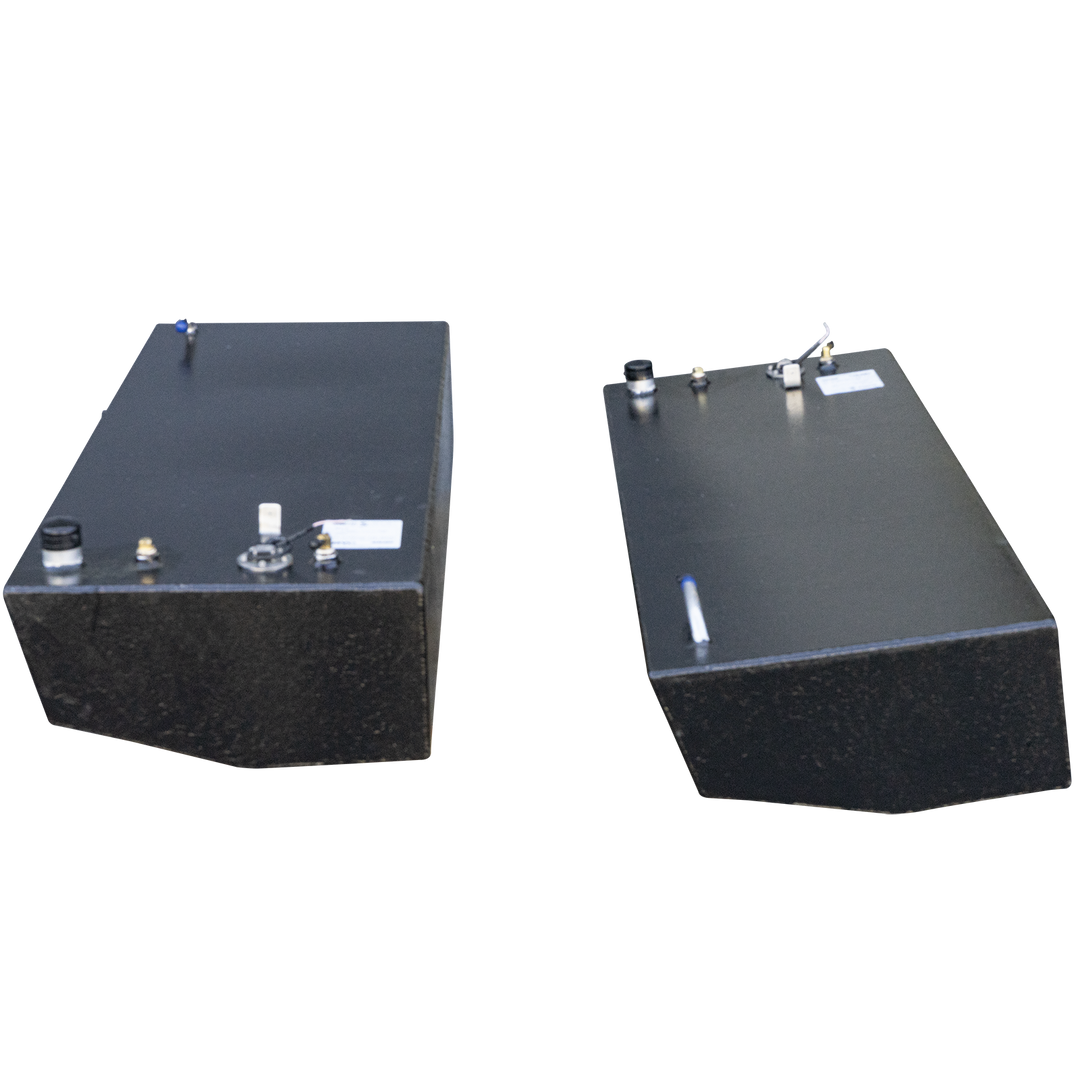 Two rectangular, dark grey fuel tanks, with various caps and fittings on their top surfaces, are positioned against a checkered black and grey background. The tanks appear to be made of metal, possibly aluminum