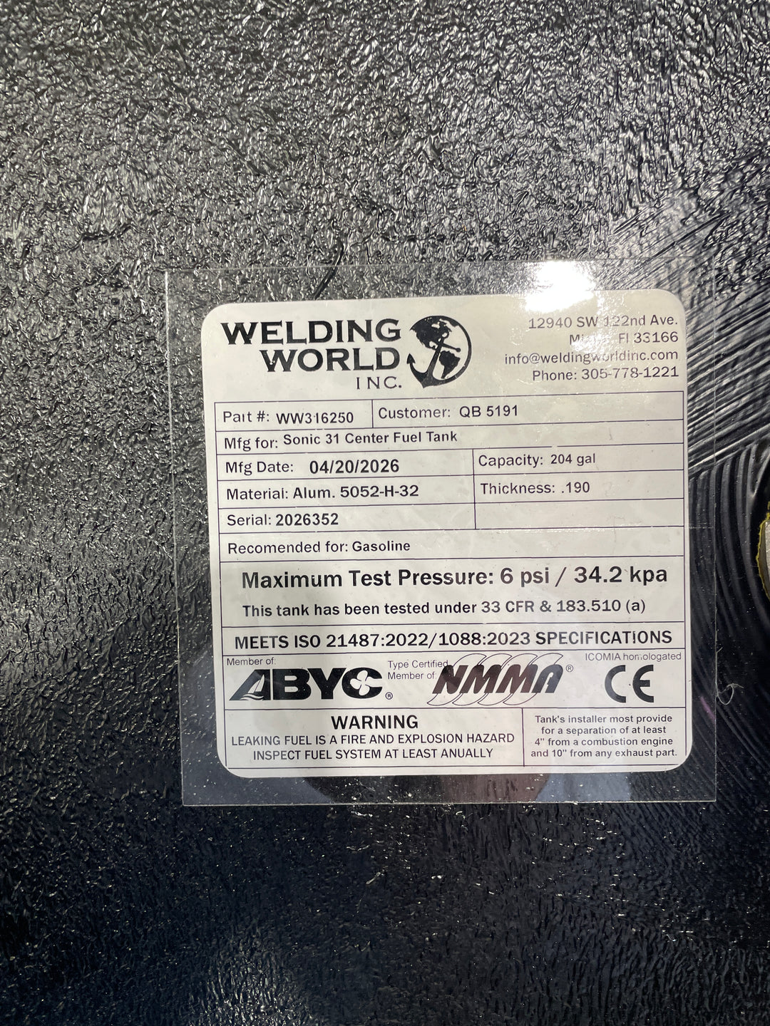Close-up photograph of the Welding World Inc. manufacturing tag for a Sonic 31 Center fuel tank replacement. The label specifies a 204-gallon capacity, heavy-duty .190" thickness 5052-H32 aluminum construction, and a 6 psi maximum test pressure. It certifies compliance with ABYC, NMMA, CE, and USCG 33 CFR § 183.510(a) safety standards, ensuring a high-quality, legally compliant OEM replacement for marine gasoline systems.