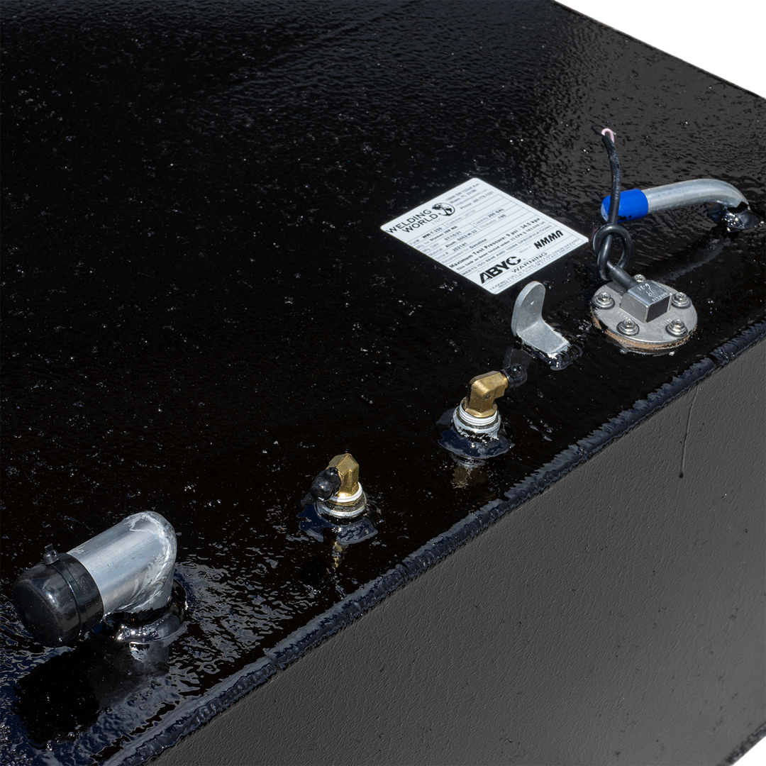A close-up view of the top of a black, textured fuel tank, showing various metallic and plastic fittings, including an L-shaped hose connector, a fuel gauge with wiring, and two smaller brass-colored fittings. A white product label from "Welding World Inc." is partially visible in the background