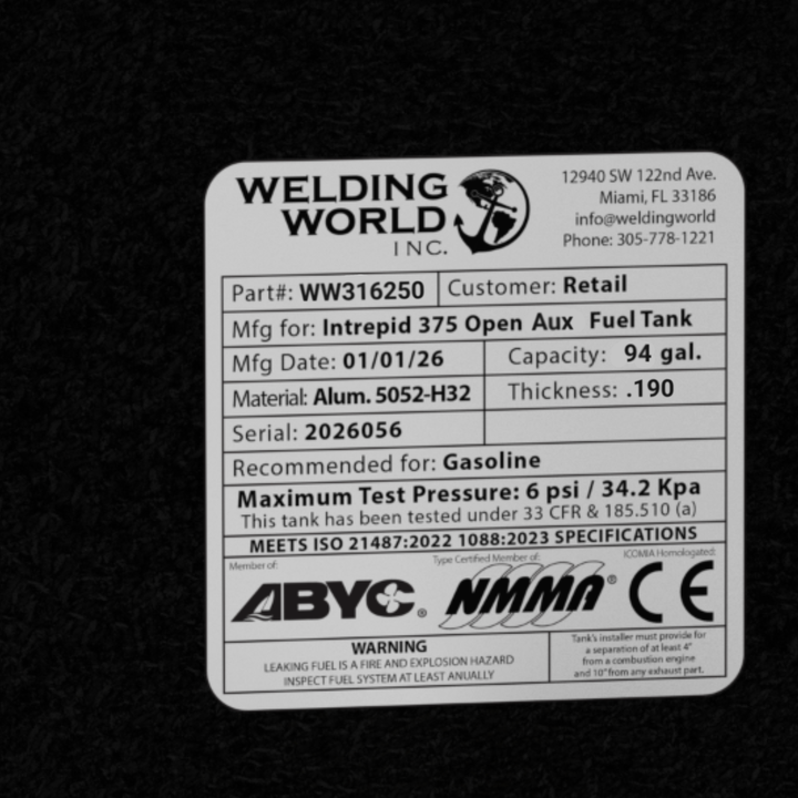 A close-up top-down photograph of the official certification label and manufacturer specification plate on a black epoxy-coated marine fuel cell for an Intrepid 375 Open Aux Fuel Tank replacement. The Welding World Inc. data plate displays critical engineering information: Part number WW316250, a 94-gallon capacity, high-grade Alum. 5052-H32 material with a .190 thickness, and a manufacturing date of 01/01/26. The label confirms the cell is recommended for gasoline with a maximum test pressure of 6 psi / 34