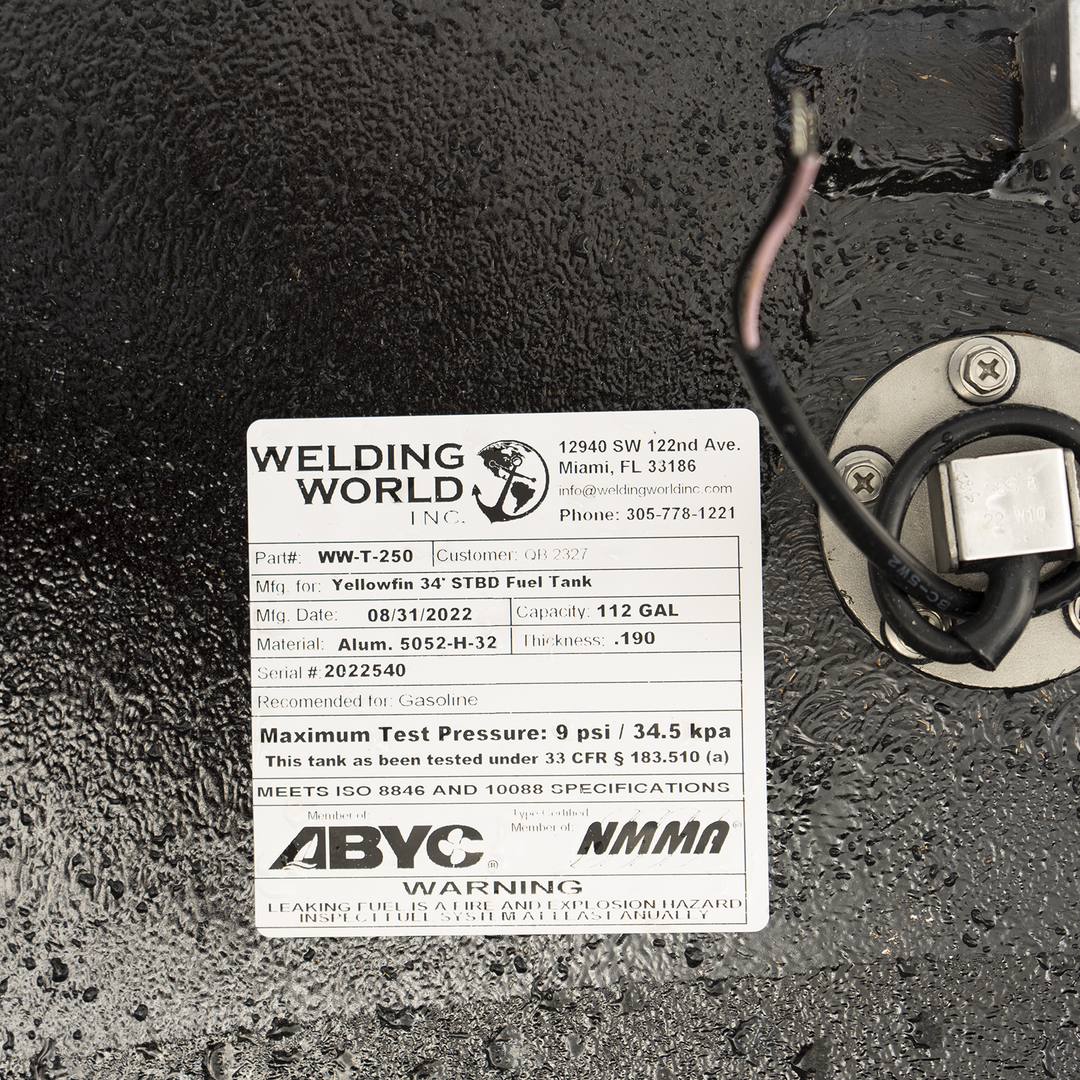 A close-up view of a product label from 'Welding World Inc.' attached to a textured black surface. The label details specifications for a 'Yellowfin 34' STBD Fuel Tank,' including Part # WW-T-250, Mfg. Date 08/31/2022, Capacity 112 GAL, Material Alum. 5052-H-32, Thickness .190, and Serial # 2022540. It also specifies the tank is recommended for Gasoline, has a Maximum Test Pressure of 9 psi / 34.5 kpa, and meets ISO 8846 and 10088 specifications. Logos for ABYC and NMMA are present