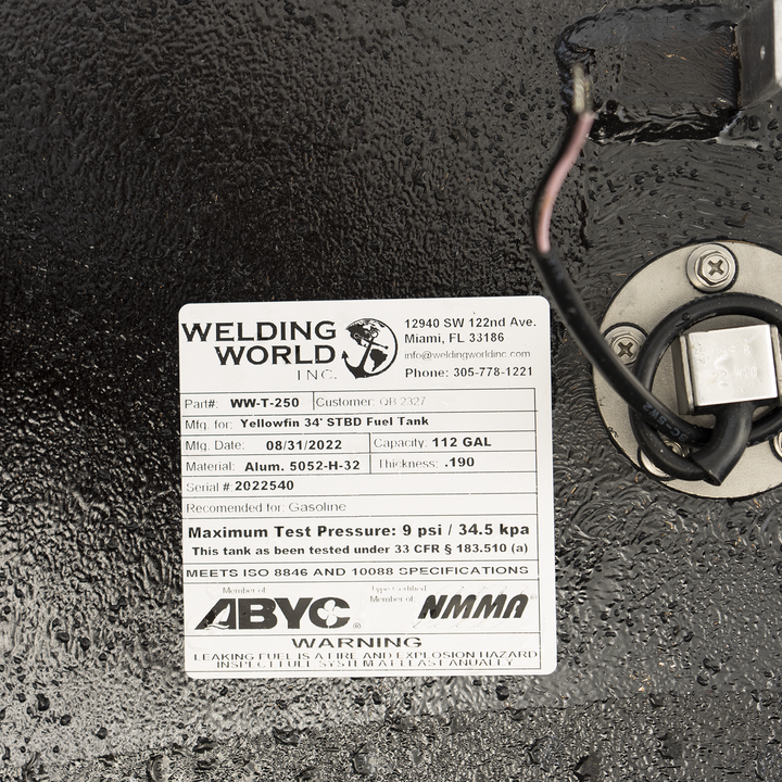 A close-up view of a product label from 'Welding World Inc.' attached to a textured black surface. The label details specifications for a 'Yellowfin 34' STBD Fuel Tank,' including Part # WW-T-250, Mfg. Date 08/31/2022, Capacity 112 GAL, Material Alum. 5052-H-32, Thickness .190, and Serial # 2022540. It also specifies the tank is recommended for Gasoline, has a Maximum Test Pressure of 9 psi / 34.5 kpa, and meets ISO 8846 and 10088 specifications. Logos for ABYC and NMMA are present